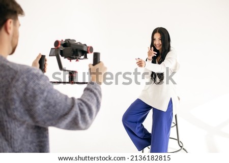 Videographer with a video camera shoots a video with a brunette girl on a white background indoors. Video blog for social media or interview. Smiling woman gesturing while working at camera