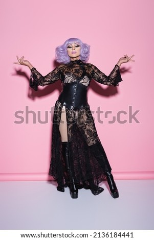 transgender person in black lace dress and violet wig standing with open arms on pink