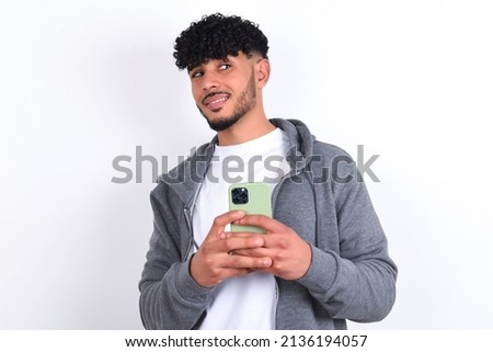 Happy young arab man with curly hair wearing casual clothes over white background listening to music with earphones using mobile phone.