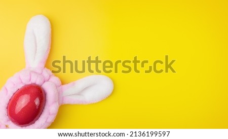 A red egg with pink rabbit ears on a yellow background. The concept of celebrating Easter.