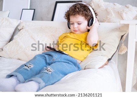 Smiling happy kid curly girl using phone in wireless headphones in bed at home