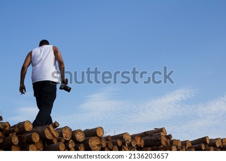 Male photographer climbing on a log pile on a blue sky day wearing white shirt, black pants and holding his camera and cigarette
