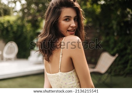 Close-up of cute caucasian young girl looking at camera over her shoulder on blurred background. Brunette with loose hair wears dress in summer. Concept of solitude with nature. Royalty-Free Stock Photo #2136215541