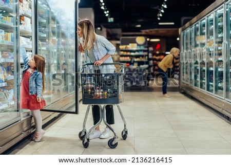 Young beautiful mother holding pushing shopping cart with her child in supermarket. Girl is choosing daily milk product picking up from shelf with her mother beside. Shopping for healthy. Royalty-Free Stock Photo #2136216415