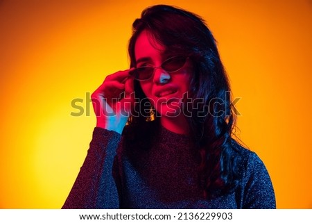 Putting eyewear on. Beautiful young excited girl with long dark hair isolated on gradient yellow background in neon light. Concept of emotions, facial expression, youth, aspiration, sales.