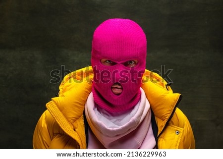 Looking at camera. Studio shot of young anonymous man wearing pink balaclava and yellow down jacket, coat isolated on dark vintage background. Concept of safety, art, fashion. Royalty-Free Stock Photo #2136229963