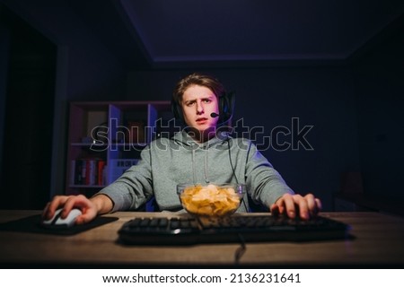 Handsome young male gamer with headset on his head with a plate of chips enjoys the computer at night at home with a serious face.