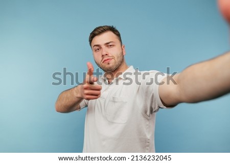Handsome man with bristles in a white T-shirt makes a selfie on a blue background with a serious face.