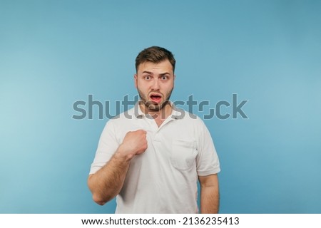 Portrait of shocked man in white t-shirt pointing finger at himself and looking unhappily at camera on blue background.