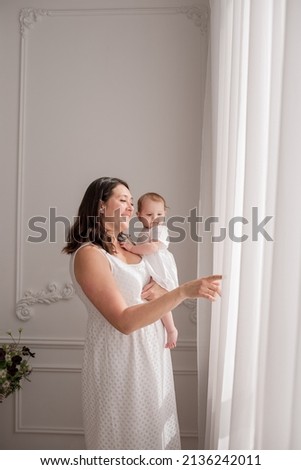 Caucasian brunette mom holds a baby girl and looks out the window in the room