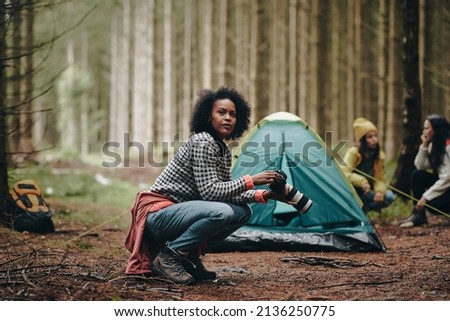 Young African American woman taking photos with a telephoto lens while camping with friends in a forest