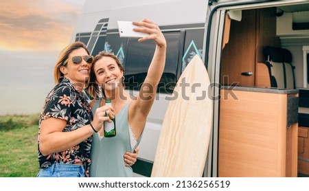 Two happy young women taking a selfie with their cell phone next to their camper van during a trip