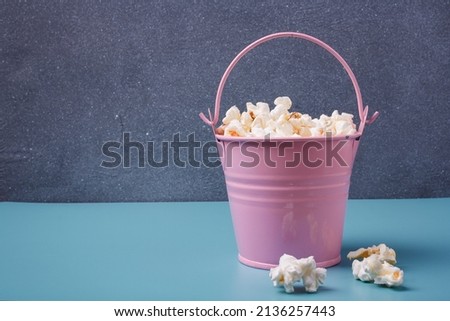 Small pink bucket of pop corn on blue and grey background. Some popcorns lying near. Copy space.