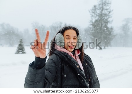 Joyful woman in warm winter clothes in the snow stands on the street and blinking shows a gesture of peace, smiling and looking into the camera. Walk in the snow.