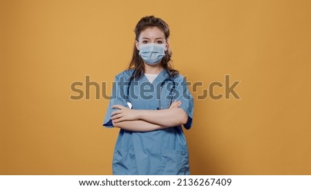 Woman doctor putting on covid mask for protection and posing confindent with arms crossed in studio with copy space background. Portrait of caucasian medic wearing protective uniform and stethoscope.