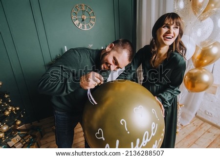 A stylish, bearded man and a cute, beautiful pregnant brunette girl are standing near a large balloon and want to pierce it with a needle