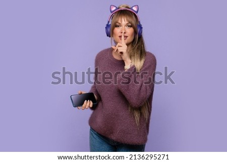 Girl in oversized sweater listens to music in wireless headphones and put index finger to lips. Woman holding cell phone and makes sign of silence. Shh... Modern technology and lifestyle concept.