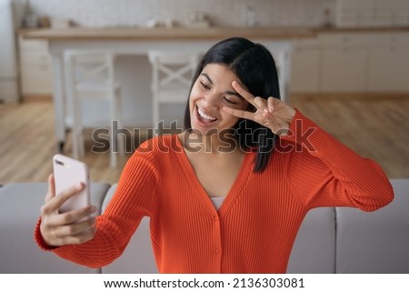Young emotional influencer using mobile phone streaming video relaxing at home. Beautiful African American woman taking selfie, showing victory sign, communication online
