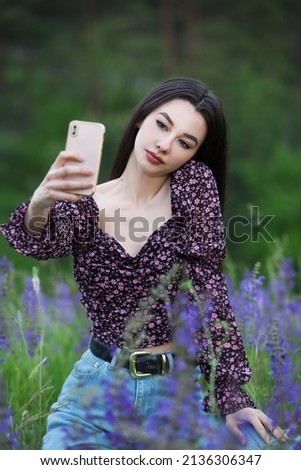 Beautiful young woman taking a selfie with a smartphone while sitting in a field with flowers.