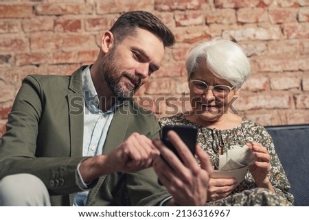 caucasian relatives son and elderly mother have a memory of past events while looking at his phone and printed pictures. High quality photo