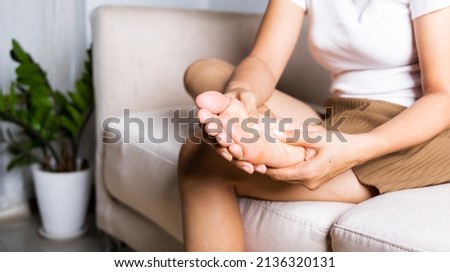 Foot pain, Asian woman sitting on sofa feeling pain in her foot at home, female suffering from feet ache use hand massage relax muscle from soles in home interior, Healthcare and podiatry medical Royalty-Free Stock Photo #2136320131