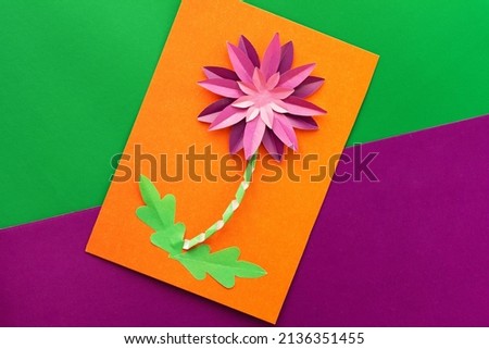 Top view of papercraft violet crysanthemum flower made by child on orange worksheet on violet and green background. Present for mother s day