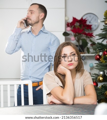 Offended young woman sitting at table at home on Christmas Eve on background with her boyfriend talking on phone