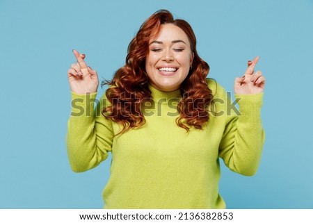 Young wistful chubby overweight plus size big fat fit woman wear green sweater waiting for special moment keep fingers crossed making wish isolated on plain blue background. People lifestyle concept
