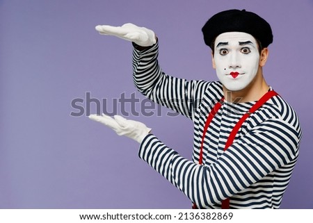 Charismatic amazing ecstatic young mime man with white face mask wears striped shirt beret hands aside like holding carrying something isolated on plain pastel light violet background studio portrait Royalty-Free Stock Photo #2136382869