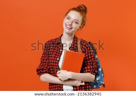 Smiling young teen redhead student girl in casual red checkered shirt backpack posing isolated on orange background. Education in high school university college concept. Mock up copy space. Hold books