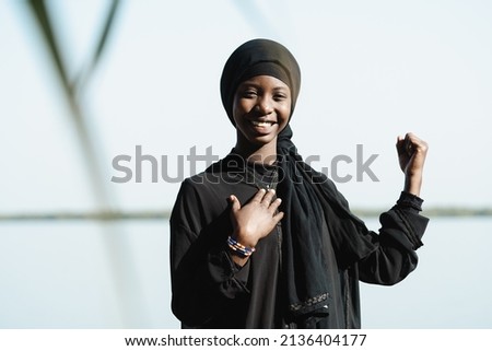 Front view of a smiling young African girl with one hand on her chest and raised fist, symbolizing the power, confidence and self-esteem of the women of her continent's future generations Royalty-Free Stock Photo #2136404177