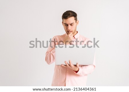 Portrait of serious pondering young man standing with laptop looking on screen with confused puzzled face on white isolated background. Studio shot of pensive male posing with pc looking at screen Royalty-Free Stock Photo #2136404361
