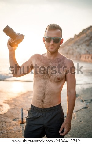 A young male photographer in shorts holds a camera in his hands stands at sunset by the sea