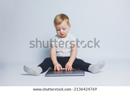 Little smart boy 3-4 years old sits with a laptop on a white background. A child in a white T-shirt and black trousers sits at a laptop and looks at the screen. Modern progressive children