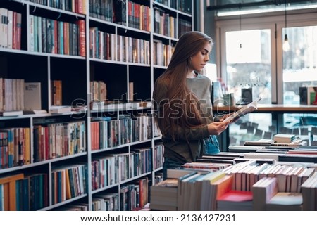 Young female customer reading a book in bookstore while buying some good literature. Woman picking a book to read. Royalty-Free Stock Photo #2136427231