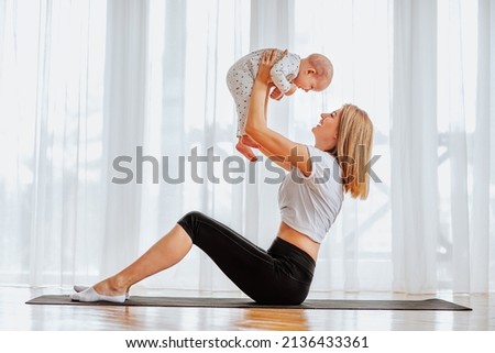 Young blond mother training at home with cute baby girl against big window. Fitness concept. Royalty-Free Stock Photo #2136433361