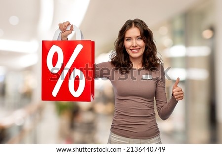 sale and business concept - happy female shop assistant or saleswoman holding shopping bag with percentage sign showing thumbs up over mall background