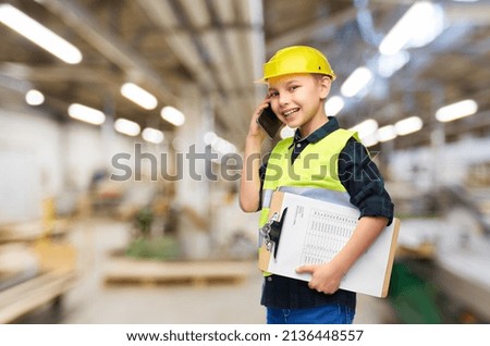 industry, manufacture and profession concept - happy smiling little boy in protective helmet and safety vest with clipboard calling on smartphone over workshop background