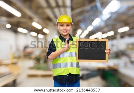 industry, manufacture and profession concept - happy smiling little boy in protective helmet and safety vest holding chalkboard over workshop background
