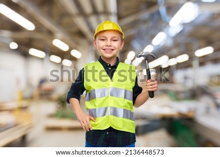 industry, manufacture and profession concept - happy smiling little boy in protective helmet and safety vest with hammer over workshop background