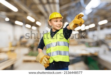 industry, manufacture and profession concept - happy smiling little boy in protective helmet, gloves and safety vest pointing finger over workshop background
