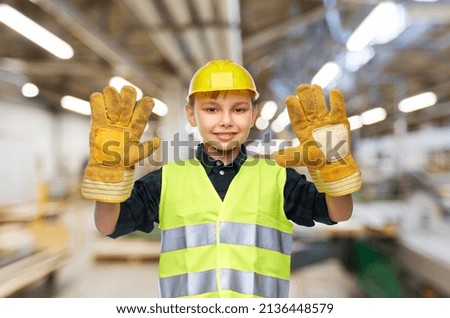 industry, manufacture and profession concept - happy smiling little boy in protective helmet and safety vest over workshop background