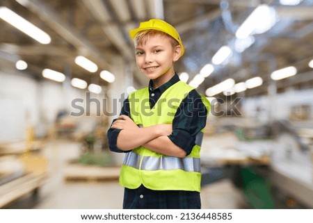 industry, manufacture and profession concept - happy smiling little boy in protective helmet and safety vest with crossed arms over workshop background