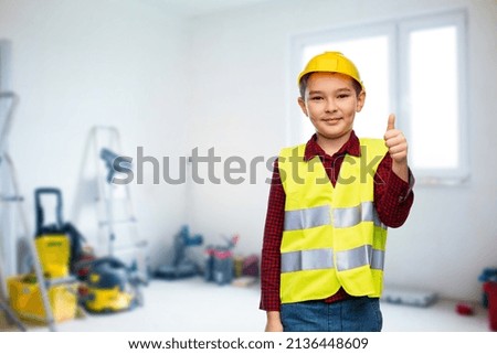 building, construction and profession concept - little boy in protective helmet and safety vest showing thumbs up over room with working equipment background
