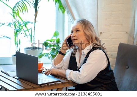 An elderly business woman is sitting in a cafe at a laptop and talking on the phone