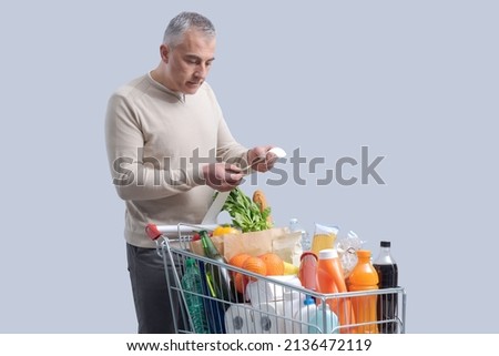 Disappointed man standing next to a full shopping cart and checking an expensive grocery bill Royalty-Free Stock Photo #2136472119
