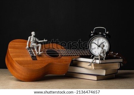 A man is sitting on a ukulele and talking to a woman sitting on a stack of books next to the clock. Composition in the dark key. Royalty-Free Stock Photo #2136474657