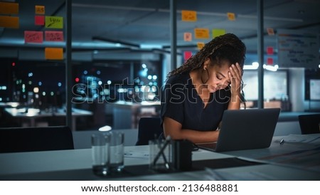 Overworked African American Businesswoman Working on Laptop Computer in Big City Office Late in the Evening. Tired Stressed Female Entrepreneur trying to Find Solution for Business Problems. Royalty-Free Stock Photo #2136488881
