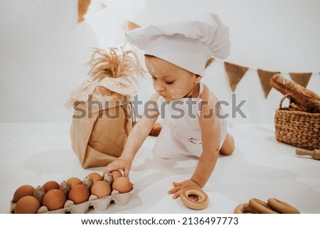 A young child in a chef's apron and hat plays the role of a baker. The table is full of clutter. The mood is sublime. There are many ingredients and different things around. Concept for bakery.