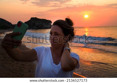 A girl with a phone takes pictures of herself against the backdrop of the sea and sunset. Red-orange sky. 